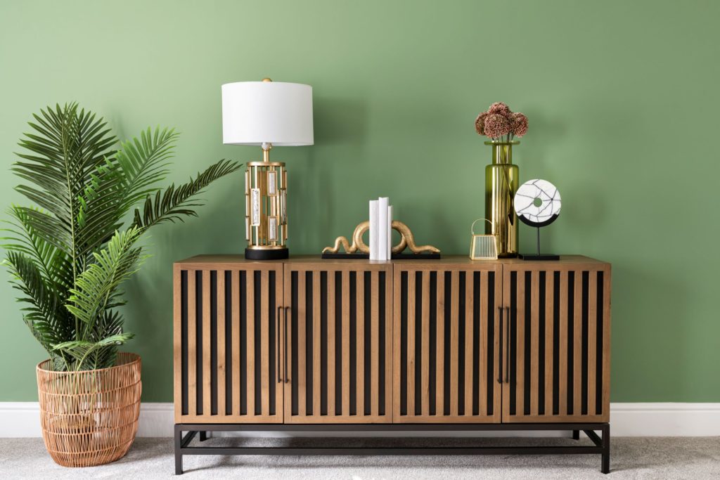 green wall with brown sideboard, gold lamp and bookends and large palm plant.