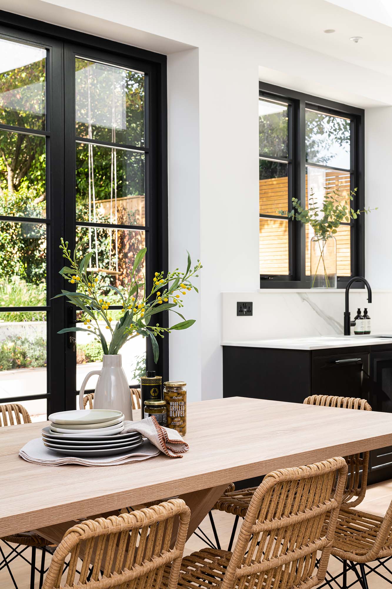 light wood dining table with wicker dining chairs and black window frames